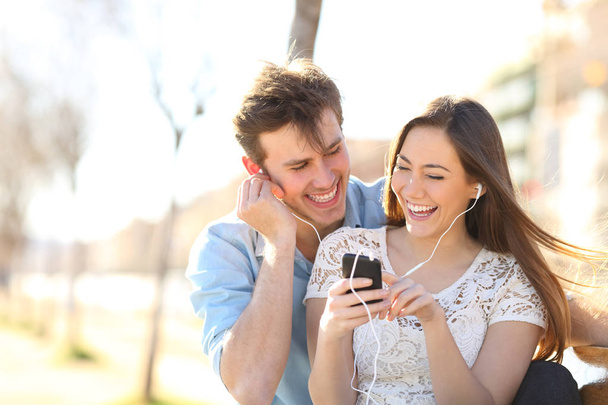 Joyful couple listening to music from a smartphone sitting on a bench in a pak - Фото, зображення