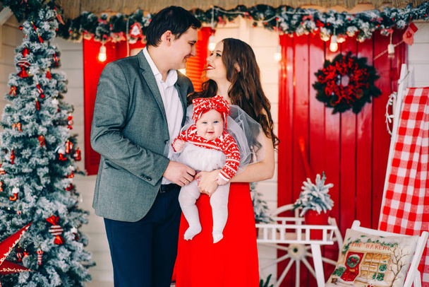 Happy parents with their little daughter in elegant clothes together in decorated holidays room with a Christmas tree and a wreath on the background. - Foto, Imagem