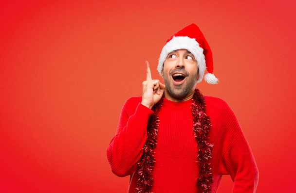 Человек в красной одежде празднует рождественские праздники стоя и думает об идее, указывая пальцем на красный фон
 - Фото, изображение