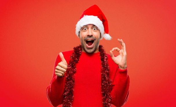 Άνθρωπος με κόκκινα ρούχα που γιορτάζει τα Χριστούγεννα διακοπές δείχνει εντάξει πινακίδα με και δίνοντας έναν αντίχειρα επάνω χειρονομία σε κόκκινο φόντο - Φωτογραφία, εικόνα