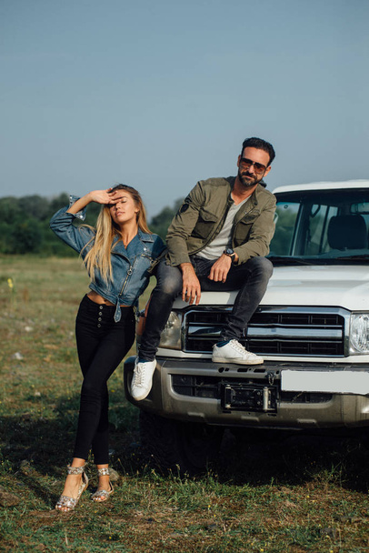 Sweet couple travel by car, road trip. Sexy young blonde and brutal man sitting on the car. Outdoor, beautiful nature landscape. - Foto, Imagem