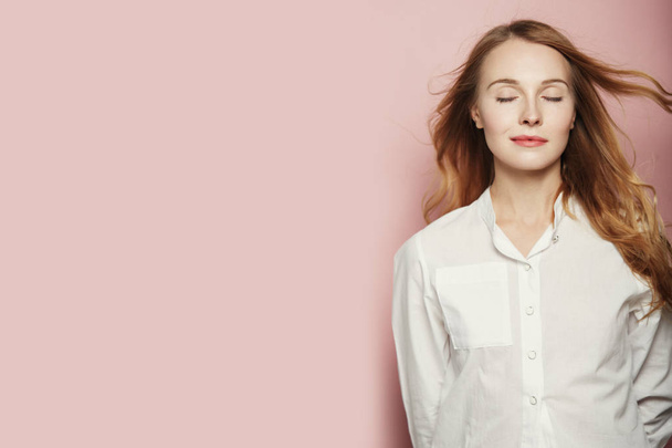 Портрет красивой белой женщины с закрытыми глазами и длинными волосами, в белой рубашке на розовом фоне
 - Фото, изображение