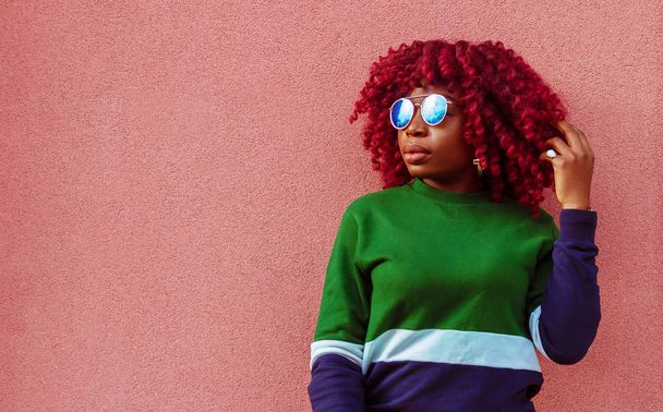 Charming young African female in blue sunglasses. Fashionable and stylish girl posing near pink wall - Φωτογραφία, εικόνα