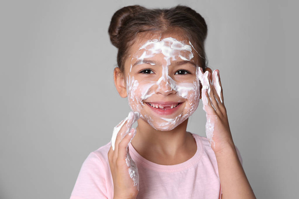 Schattig klein meisje met zeep schuim op gezicht tegen de grijze achtergrond - Foto, afbeelding
