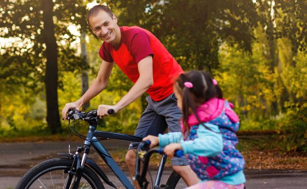 χαρούμενος νεαρός πατέρας και η μικρή κόρη του ιππασία ποδήλατα σε καταπράσινο πάρκο - Φωτογραφία, εικόνα