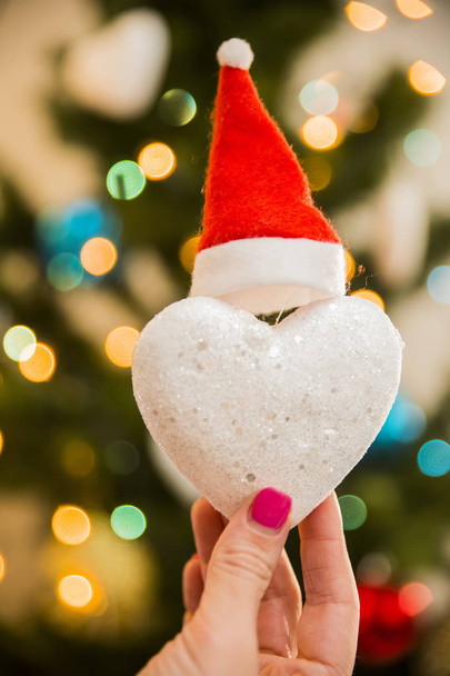 Juguete de Navidad de corazón de nieve vestido con un sombrero de Navidad en la mano sobre el fondo del árbol de Navidad
 - Foto, imagen