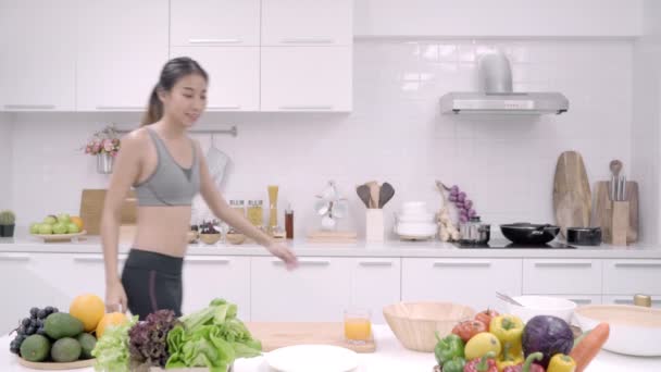 Nuori aasialainen nainen juo appelsiinimehua keittiössä, kaunis nainen urheiluvaatteissa käyttää orgaanisia hedelmiä paljon ravintoa tekemällä appelsiinimehua itse kotona. Terveellisen ruoan käsite
. - Materiaali, video