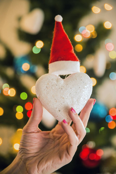 Weihnachtsspielzeug aus Schnee Herz in einem Weihnachtsmütze in der Hand auf dem Hintergrund des Weihnachtsbaums gekleidet - Foto, Bild