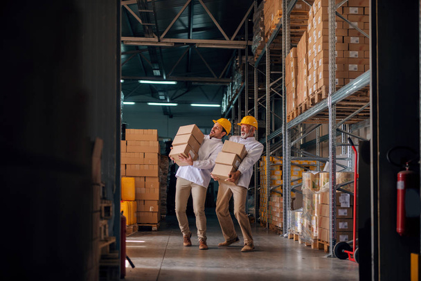 Compañeros de trabajo llevando cajas con cascos en las cabezas. Almacenamiento interior
. - Foto, Imagen