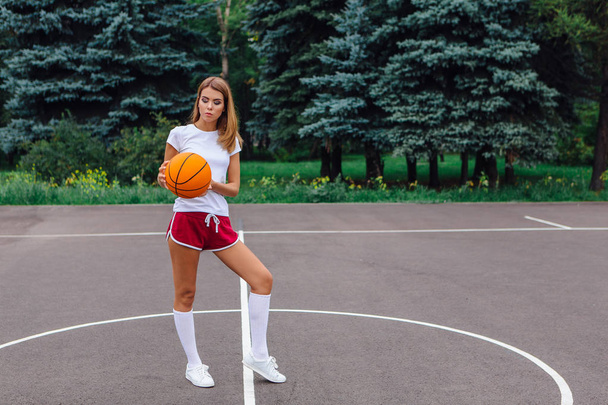 Beautiful young blonde girl dressed in white t-shirt, shorts and sneakers, plays with ball on a basketball court outdoors. Copy space. - Фото, зображення