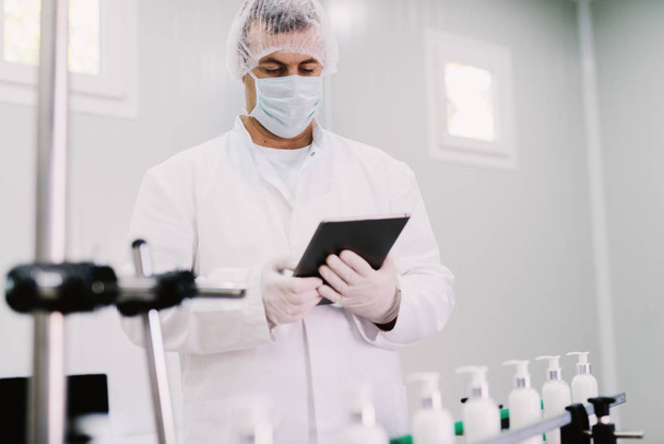 Bild eines jungen Mannes in steriler Kleidung mit Tablette. Steuerung der Produktionslinie ab Werk. Flaschen mit kosmetischen Produkten am Fließband. - Foto, Bild