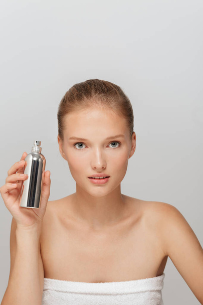 Portret van schitterend meisje zonder make-up holding parfumfles in hand zorgvuldig in de camera kijkt uit over de grijze achtergrond - Foto, afbeelding