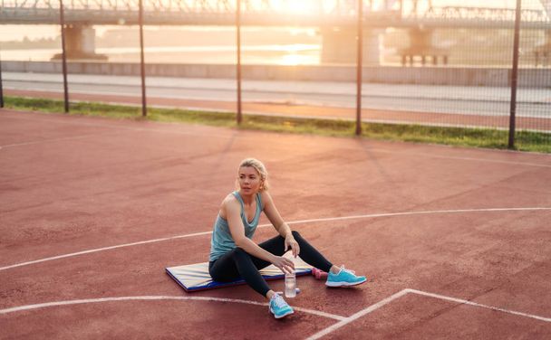 Moe jonge sportieve leuk blond meisje zitten buiten op de mat van een opleiding na een harde training. Vroege ochtend training. - Foto, afbeelding