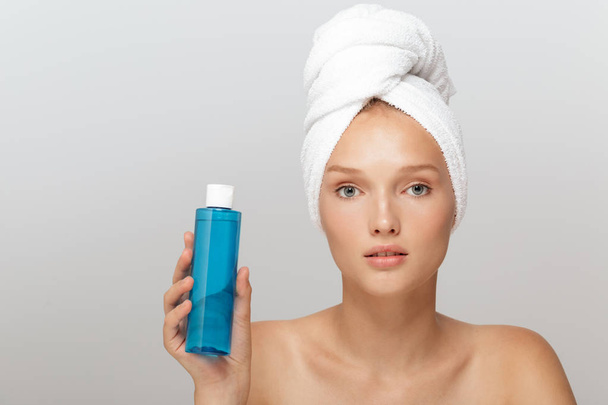 Portrait of young beautiful woman without makeup with white towel on head holding blue cosmetic bottle in hand thoughtfully looking in camera over gray background - Photo, Image