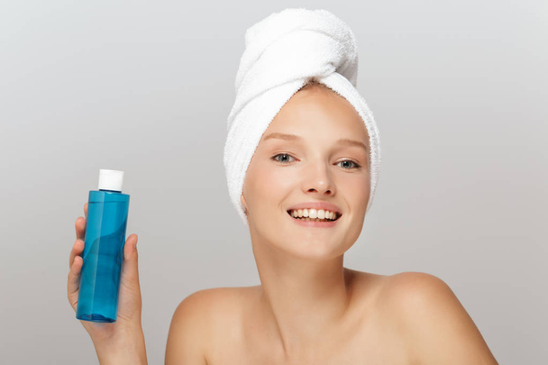 Portret uśmiechnięte dziewczyny bez makijażu z biały ręcznik na głowę trzyma niebieski butelki kosmetyczne w ręce radośnie w aparacie patrząc się na szary tło - Zdjęcie, obraz
