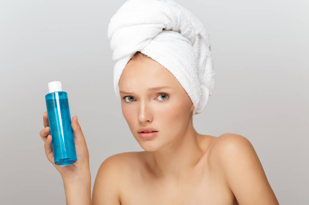 Portrait of young upset woman without makeup with white towel on head holding blue cosmetic bottle in hand thoughtfully looking in camera over gray background - Photo, Image