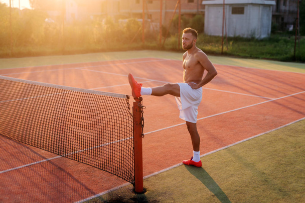 Ισχυρό σπορ άνδρα τεντώνοντας τα πόδια του πριν από την κατάρτιση. Στέκεται στο τένις παιδική χαρά το πρωί ζεστό καλοκαίρι. - Φωτογραφία, εικόνα