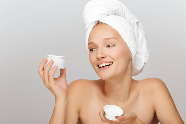 Portret van vrolijk meisje zonder make-up met witte handdoek op hoofd houden gezicht crème in hand gelukkig opzij kijkt uit over de grijze achtergrond - Foto, afbeelding