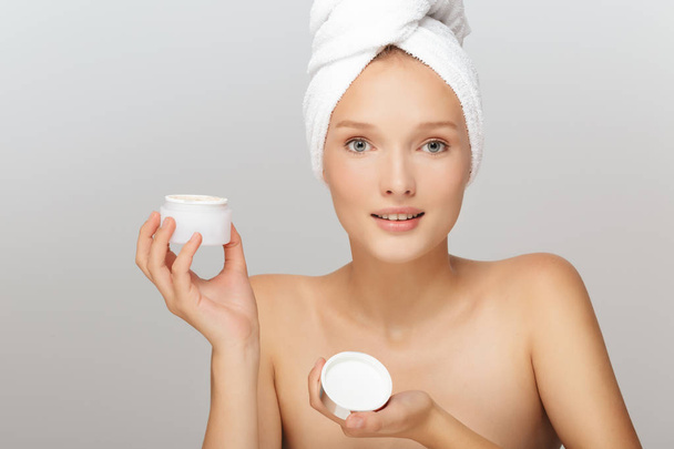 Portrait of pretty girl without makeup with white towel on head holding face cream in hand dreamily looking in camera over gray background - Фото, изображение