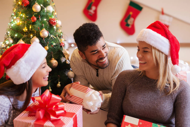 Zblízka happy přátelé drží dary při sezení v ložnici. V pozadí vánoční dekorace. Vánoční svátky koncept. - Fotografie, Obrázek