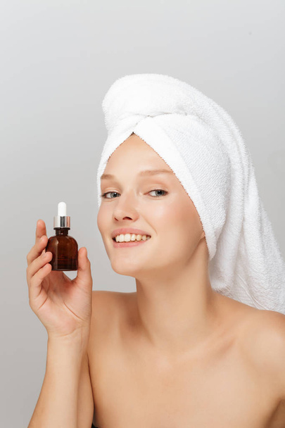 Portret atrakcyjna Pani bez makijażu z biały ręcznik na głowę, trzymając pojemnik płynu w ręce szczęśliwie w aparacie patrząc się na szary tło - Zdjęcie, obraz