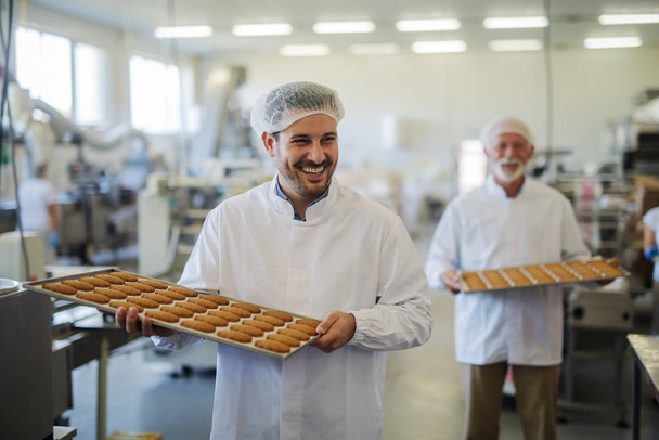 Photo de deux employés masculins souriants heureux en vêtements stériles portant des plateaux remplis de biscuits frais dans l'usine alimentaire. S'entraider et avoir l'air joyeux
. - Photo, image