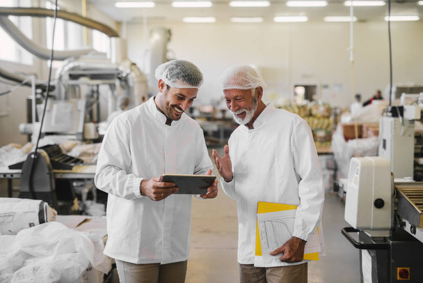 Dos alegres colegas masculinos en ropa estéril de pie en la fábrica de alimentos y mirando los documentos de venta en la tableta digital
. - Foto, imagen