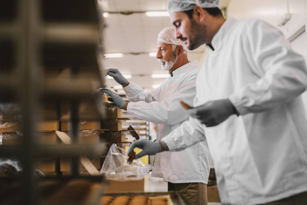 Imagen de dos empleados de la fábrica de alimentos masculinos en ropa estéril empaquetando galletas recién hechas. De pie en habitación luminosa
. - Foto, Imagen