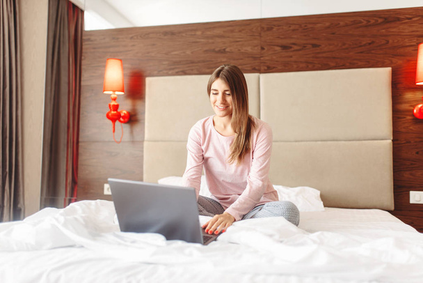 Femme souriante assise au lit et utilise un ordinateur portable, bonjour, chambre interoir sur fond
 - Photo, image