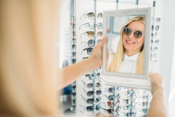 Cliente fêmea dispara óculos de sol na loja de óptica. Seleção de proteção de olhos na loja de óculos, conceito de optometria
 - Foto, Imagem