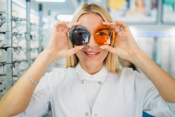 Женщина наносит на глаза линзы разных цветов, демонстрируя очки в магазине оптики. Выбор очков с профессиональным оптометристом, выбор солнцезащитных очков, концепция eyecare
 - Фото, изображение