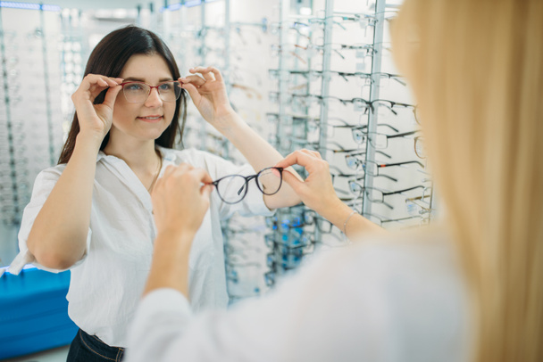 Female optician and buyer chooses glasses frame against showcase with eyeglasses in optics store. Selection of spectacles with professional optometrist - Photo, Image