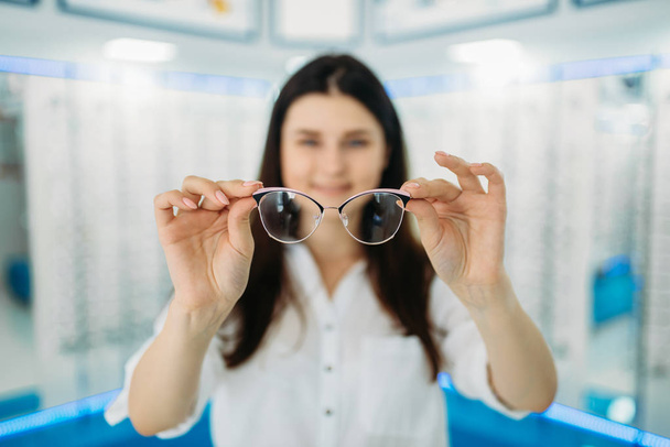 Young female customer holds glasses in hand, optic store, showcase with spectacles on background. Professional eye care, eyeglasses choice - Photo, Image