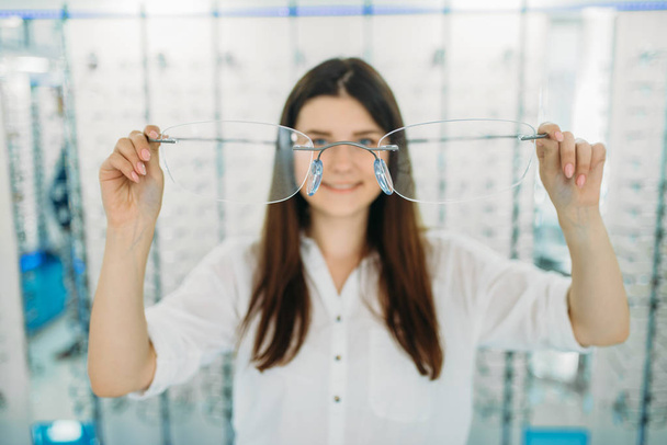 Young woman holds huge decorative glasses, optic store, showcase with spectacles on background. Professional eye care, eyeglasses choice - Foto, imagen