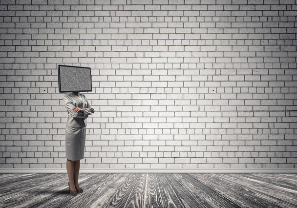 Business woman in suit with TV instead of head keeping arms crossed while standing inside empty room with gray brick wall on background. - Foto, Imagen