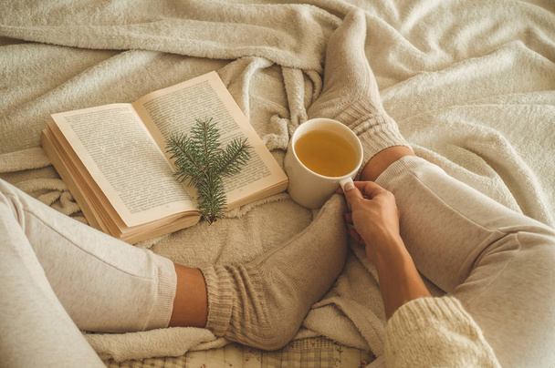 Acogedora noche de invierno, cálidos calcetines de lana. La mujer está acostada con los pies sobre una manta blanca y peluda y leyendo un libro. Acogedora escena de ocio
 - Foto, Imagen