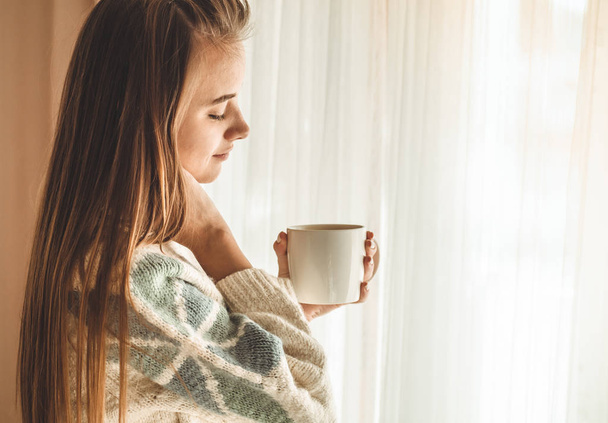 Cozy home. Woman with cup of hot drink  by the window. Looking at window and drink tea. Good morning with tea. Young girl relaxing - Foto, Bild