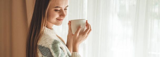 Ζεστό, άνετο σπίτι. Γυναίκα με φλιτζάνι ζεστό ρόφημα από το παράθυρο. Κοιτάζοντας από το παράθυρο και να πιείτε καφέ. Καλημέρα με τσάι. Νεαρό κορίτσι χαλάρωση - Φωτογραφία, εικόνα