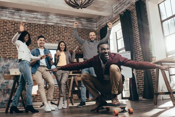 group of happy multiethnic coworkers having fun with skateboard in loft office - Photo, Image