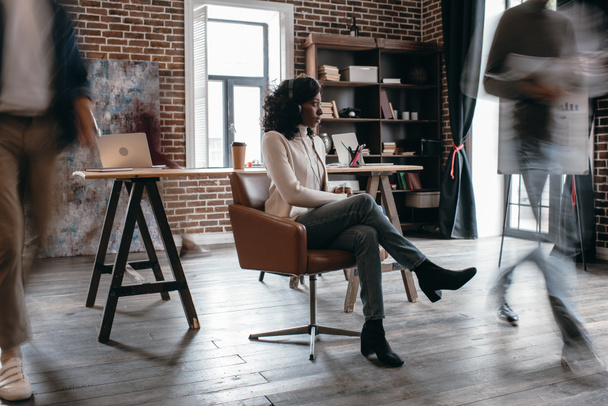 african american businesswoman in heaphones sitting on chair with colleagues in motion blur in modern loft office - Foto, Imagem