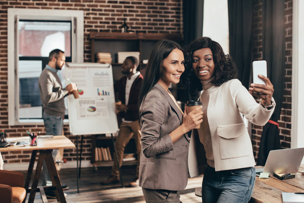 multiethnic couple of business women taking selfie on smartphone in loft office with colleagues on background - Photo, Image