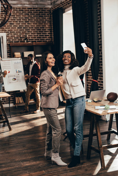 smiling multiethnic couple of business women taking selfie on smartphone in loft office with colleagues on background - Фото, изображение