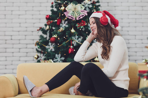 Ein junges Paar mit rotem Hut sitzt auf einem Sofa zwischen Weihnachtsbäumen und hört Musik mit Kopfhörer vor einem riesigen Weihnachtsbaum. Konzept für gute Zeit und Glück an Weihnachten. - Foto, Bild