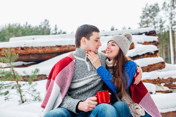 Young loving couple dressed in blue sweater sitting with red coffee cups on the firewood in winter forest - Φωτογραφία, εικόνα