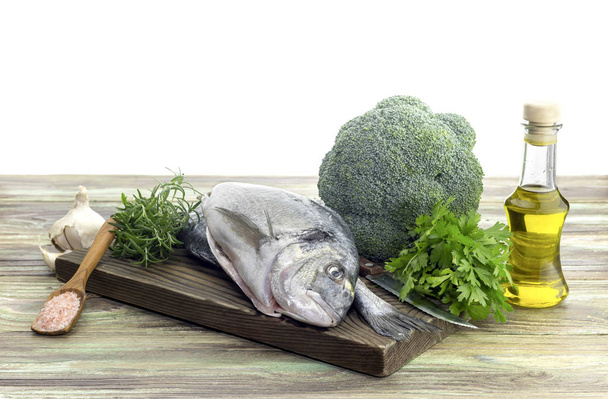 Sargos-do-mar de peixe (Sparus aurata) em uma tábua de corte, azeite, brócolis, alho e ervas aromáticas em uma mesa de madeira close-up
. - Foto, Imagem