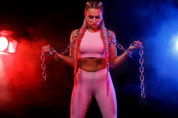 Sporty fit woman dancer and athlete with chains makes fitness exercising on black background with colorful lights. - Zdjęcie, obraz