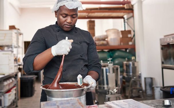 Worker standing at a table in an artisanal chocolate making factory mixing melted chocolate in a bain marie with a spoon - 写真・画像