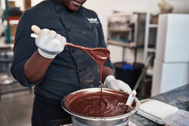 Close-up van een werknemer een lepel gesmolten chocolade vol te houden tijdens het mixen van de ingrediënten in een ambachtelijke chocolade maken van fabriek - Foto, afbeelding