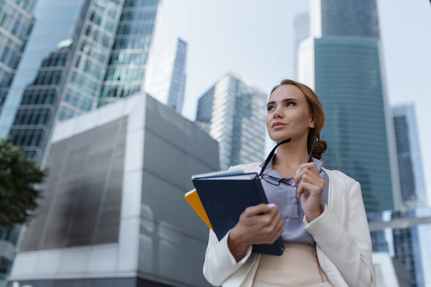 Young woman with folders and documents in her hands standing among the skyscrapers of the modern business center - Photo, Image