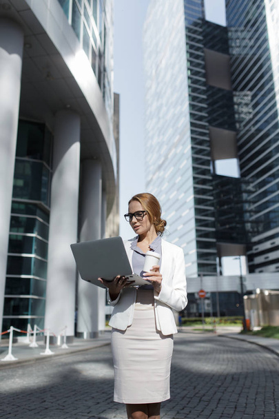 Young beautiful business woman with cup of coffee and laptop in her hands standing among the downtown - Foto, imagen